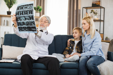 Mature aged attractive high-skilled caring male doctor explaining results of x-ray MRI image to his sick child patient and his mom during visiting their at home. Computed tomography.の写真素材