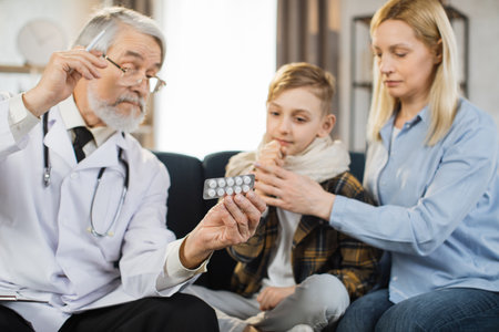 Mature caucasian male doctor giving green blister with tablets for sick preschool coughing boy patient, sitting with his blond middle age mother on the sofa in living room, during home visit.の写真素材