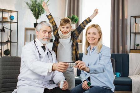 Pleasant mature male doctor , talking with concentrated pretty caucasian woman about taking of medicines during home visit of sick boy, standing with raised hands rejoicing in health and recovery.の写真素材