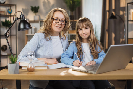 Portrait of joyful small daughter and her middle age mother doing homework and looking at camera. Young mom and her cute girl happy to make right tasks, sitting at home.の写真素材