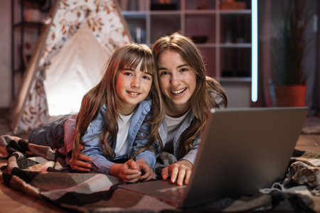 Beautiful cute sisters or friends lying on floor embrace outside wigwam using laptop computer. Happy girls spending evening time together watching favorite movie on digital device at home.の写真素材