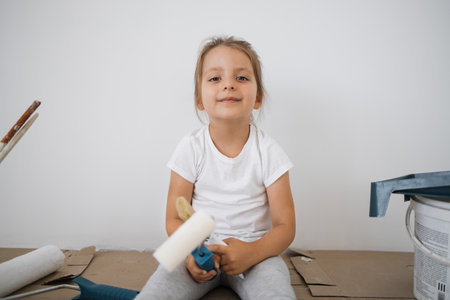 Little blonde cute girl with wall paint roller sitting on the floor during home renovation on white wall background.の写真素材