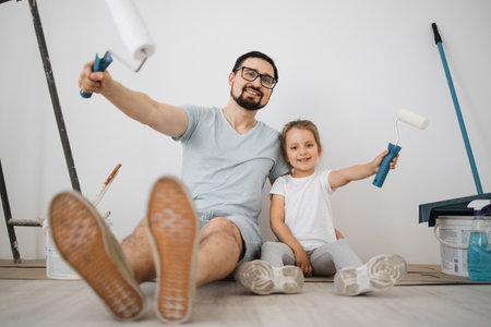 Cheerful caucasian man father and his cute little girl daughter sitting on floor in white light room background with paint rollers in their hands.の写真素材