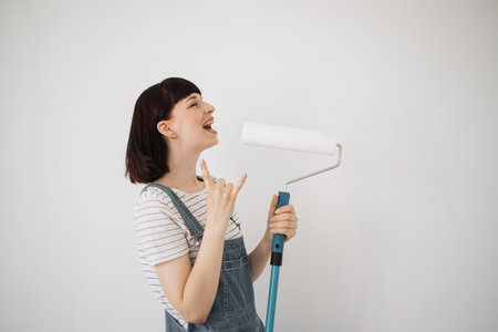 Young beautiful woman with red hair singing song at new home, girl using into a mop and a paint roller instead of real microphone showing rock signの写真素材