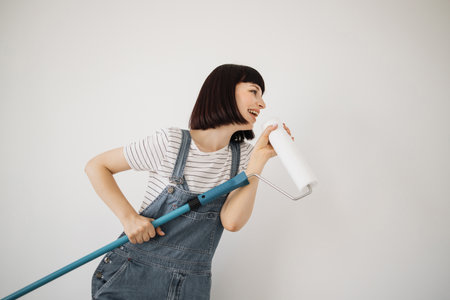 Happy cute caucasian female singing song using roller brush as a microphone. Young woman having fun during home repair and renovationの写真素材