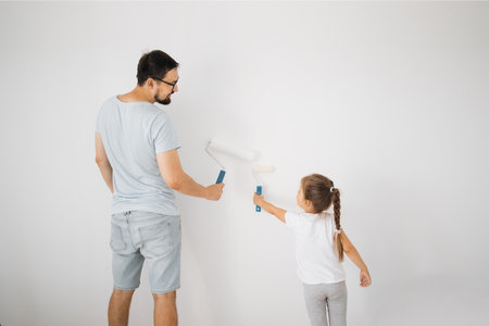 Repair in the apartment. Back view of happy family of father and little cute child daughter paints the wall with white paint.の写真素材