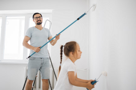 Young family of attractive father and cute little daughter having fun together while repairing room. Little girl with paint roller coloring with daddy the wall. Focus on dad.の写真素材