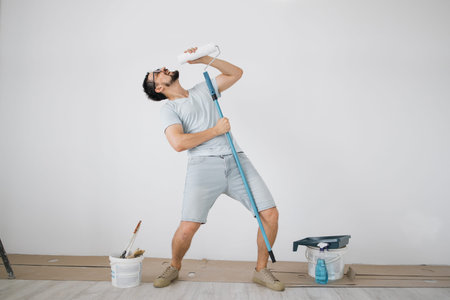 Happy caucasian male singing song using roller brush as a microphone. Young man having fun during...