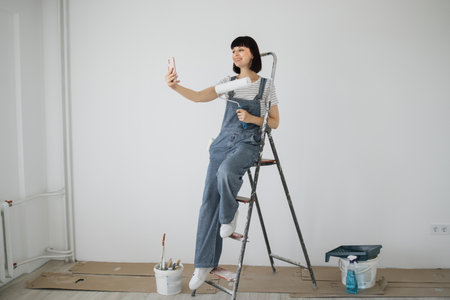 Red hair girl with cute smile holds a paint roller and smart phone in her hands. Young woman leaning on a ladder that she uses during repairs and having video call on her phone.の写真素材