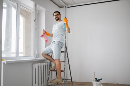Happy caucasian man professional window cleaner cleaning with equipment and liquid soaps apartment...