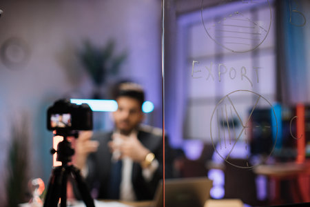 Focus on glass board with graphs and charts of export and trader or office worker sitting in front of video camera in blurred background in evening office.の写真素材