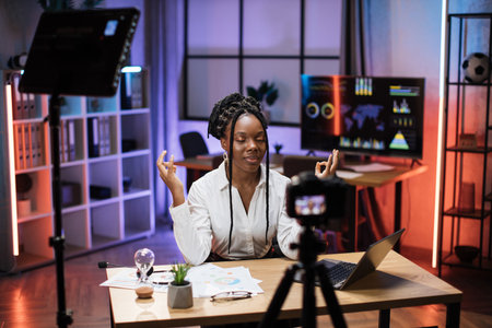 Relaxed motivated african american businesswoman in formal wear meditating with closed eyes and mudra hands in front of camera during recording video for business vlog in evening officeの写真素材