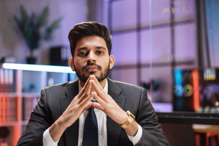 Indian bearded man in formal suit sitting at desk with modern laptop and looking at camera with...