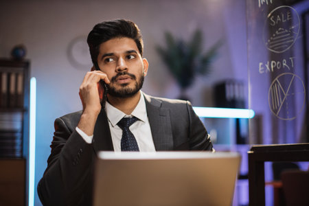 Close up portrait of handsome concentrated hardworking bearded indian businessman or manager in suit having business conversation with smart phone and laptop computer in evening office.の写真素材