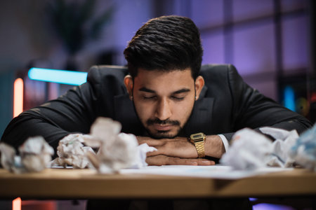 Portrait of tired overworked bearded arab businessman in formal suit, sleeping on desk with sheets of crumpled papers in eveninng office.の写真素材