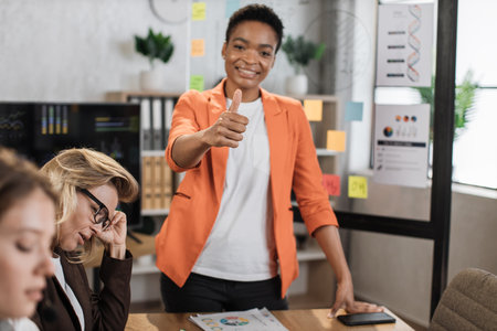 Young african woman standing at boardroom showing thumb up and looking at camera with confidence in eyes. Multi ethnic office workers talking while sitting at table at office.の写真素材