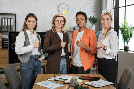 Portrait of female economists in formal wear looking at camera while standing together at office with graphs and charts. Multi ethnic brokers spending time for examining trade market showing thumb upの写真素材