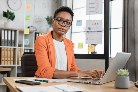 Attractive young african woman with dark hair in eyeglasses looking at camera with confidence in eyes while sitting at table with papers and laptop. Concept of success, career and technology.の写真素材