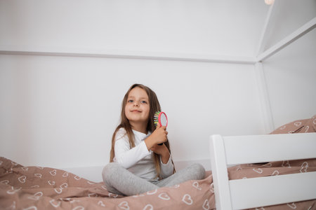 Close up portrait of sweet little girl brushing her long hair and smiling while sitting on her scandinavian wooden house frame bed at home.の写真素材