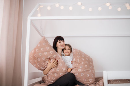 Satisfied caucasian young lady embracing preschooler girl after fighting with pillows, sitting on...