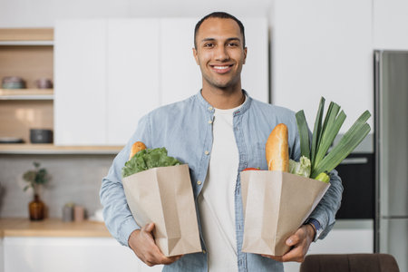 Young attractive indian man in kitchen holding two paper grocery shopping bags full of fresh healthy...