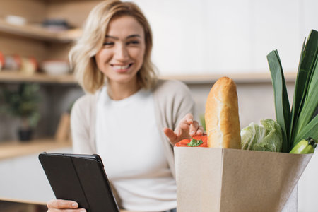 Focus on hand holding tablet and paper bag with vegetables. Happy woman cooking healthy food looking on paper bag with vegetables while reading recipe on digital tablet in modern kitchen at home.の写真素材