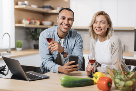 Multiracial couple using smartphone while having breakfast at kitchen, positive young man and blond...