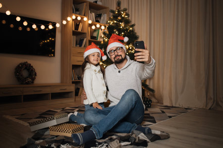 Young happy father and little cute daughter in santa hats having video call or doing selfie while sitting with smart phone in front of decorating Christmas tree in living room.の写真素材