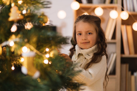 Little adorable girl in knitted sweater is hanging decoration on Christmas tree with light. Child decorating Xmas tree in beautiful living room. Happy family holiday New Years concept.の写真素材