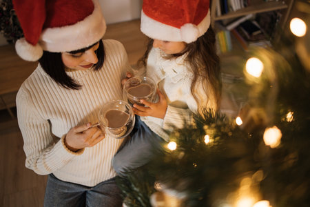 Cropped top view portrait of beautiful young caucasian mother and her cute little daughter in santa hat drinking hot cocoa or chocolate from cups near decorated Chritmas tree.の写真素材