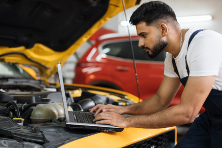 Young man car mechanic maintains vehicle using diagnostic laptop computer for checking engine problems, modern technology in the car repair shop.の写真素材