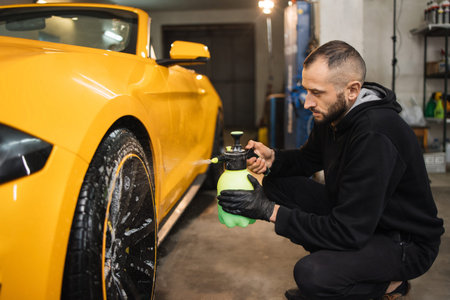 Side view of handsome bearded male car wash worker holding in hand special chemical cleansing solution and spraying it on car wheel rim. Car wash and detailing concept.の写真素材
