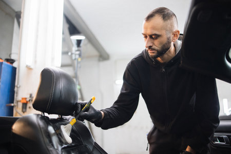 Close up horizontal portrait of young handsome male worker in black uniform and gloves, cleaning car front seat with special foam and soft brush. Car detailing, disinfection, cleaning.の写真素材