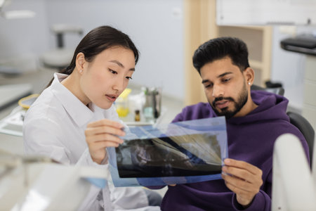 Focus on x ray picture, attractive young bearded man, sitting on dentist chair and looking at x-ray scan image of teeth together with his cheerful female asian dentist at clinic.の写真素材