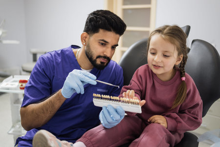Young professional bearded male dentist choosing color of filling from palette for little female...