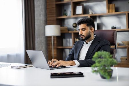 Young bearded businessman typing on laptop during work in office. Concentrated adult successful man wearing official suit sitting at wooden desk indoor.の写真素材