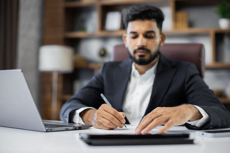 Focus on hands, young bearded male accountant working from modern office using laptop while holding a document and pen in his hands and sign new deal or writing report.の写真素材