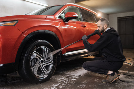 Car wheel, rim or alloy, washing. Car cleaning with water jet. Horizontal shot of pleasant young guy in white t-shirt and overalls, washing red car wheel with high pressure water jet.の写真素材