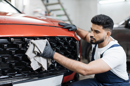Car detailing, wash and cleaning concept. Young bearded male professional car wash worker in black rubber gloves, holding the gray microfiber and wiping grille of red luxury car.の写真素材