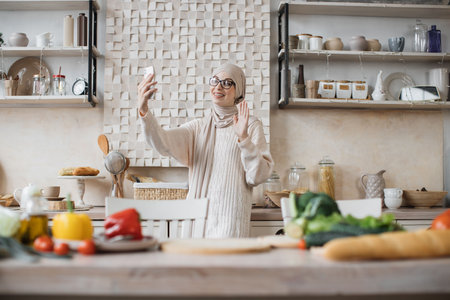 Pretty young arabian woman preparing dinner searching vegetable recipes diet menu cookbook app using smartphone Smiling muslim female doing selfie or having video call while cooking healthy vegan foodの写真素材