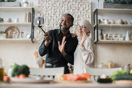 Multiethnic family having video call using selfie stick while providing cooking online class healthy vegan food Happy young couple blogger showing to followers how prepare dinner with fresh vegetableの写真素材