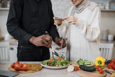 Closeup of hands of african man holding spoon and fork, mixing ingredients in bowl, preparing delicious healthy salad, meal on table dinner in kitchen, next to his wife taking photos for social mediaの写真素材