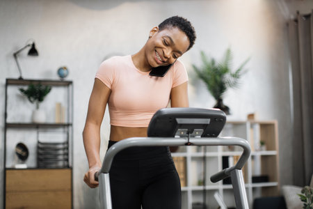 Front view of african sporty woman in pink t-shirt working out in treadmill while having phone call...