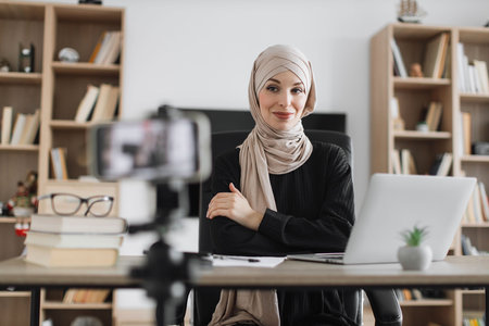 Beautiful muslim woman in hijab talking while looking at and recording video on modern smartphone. Female blogger sitting at home and doing live stream.の写真素材