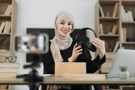Arab woman recording video on phone camera while unpacking box with new wireless headphones. Female influencer sharing with subscribers her positive feedback about new order.の写真素材