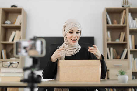 Excited smiling muslim woman in casual wear and hijab recording video on camera while unpacking gift boxes. Female blogger sharing her emotions with her subscribers in social networks.の写真素材