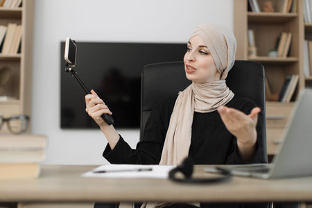 Attractive young muslim woman in hijab and headset taking selfie on her smart phone using stick. Female blogger sitting at home and doing live stream.の写真素材
