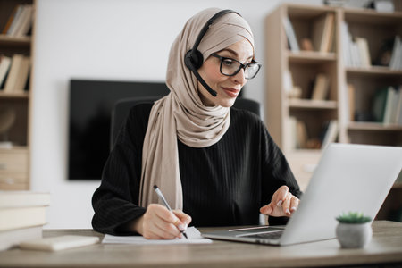 Attractive confident muslim business woman, office manager, wearing headset and hijab using laptop while making, writing financial report, using pen, on paper working indoors.の写真素材