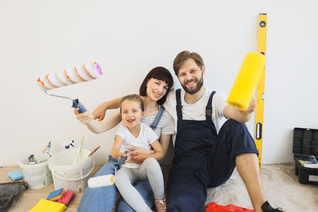 Cheerful caucasian family of father, mother and cute little girl daughter sitting on floor in white light room background with paint rollers in their hands.の写真素材