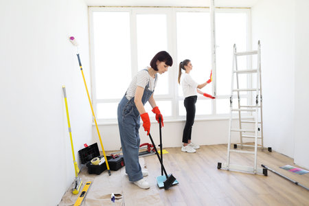 Young beautiful women cleaning up new apartment after renovation. One girl sweeping floors in house with broom and dustpan, and another wiping the window.の写真素材